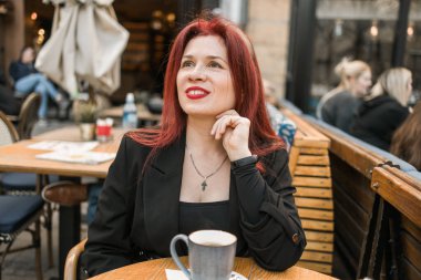 Restoranın terasında parlak kırmızı rujlu güzel bir kadın oturuyor. Kahvede poz veren siyah elbiseli kızıl saçlı harika bir kadın.