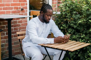 Kültürel ve etnik kıyafetleri olan Afrikalı Amerikalı hippi dışarıda akıllı telefondan sohbet ediyor sosyal ağlarda arkadaşlarıyla mesajlaşıyor. Milenyum ve İnternet kavramı.