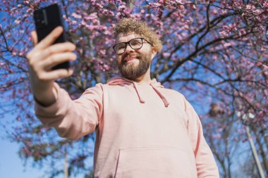 Mutlu kıvırcık adam internetteki iletişimi için çiçek açan ağacın arka planında selfie çekiyor. Hafta sonu ve sosyal ağlar