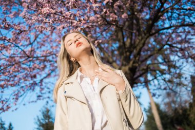 Şık takım elbiseli, güzel, kıvırcık saçlı, güzel bir kadının çiçek açan kiraz ağacıyla poz verdiği moda fotoğrafı. Reklam metni için boşluk ve boşluk kopyala.