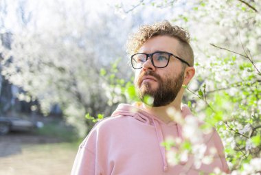 Sakura çiçeklerinin ya da çiçek açan bahar ağacının yanında sakallı ve bıyıklı bir adam. Yumuşak ve nazik bir kavram. Arka planda çiçekleri olan şık saç kesimi olan sakallı adam, yaklaş. Yakınlarda bir hippi