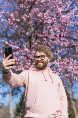 Mutlu kıvırcık adam internetteki iletişimi için çiçek açan ağacın arka planında selfie çekiyor. Hafta sonu ve sosyal ağlar