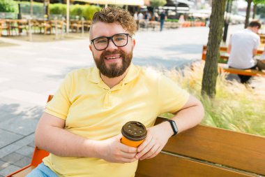 Millennial gülümseyen sarı polo tişörtlü gözlüklü öğrenci kahve içer, kahvaltı yapar, bahar güneşli yeşil şehir parkında dinlenir. Şehir eğlencesi konsepti