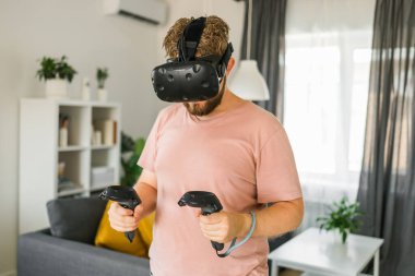 Sanal gerçeklik kulaklığı takan ve evdeki bir video oyununda kumanda tutan enerjik genç bir adam. VR macera video oyunu oynayan aktif adam oturma odasında