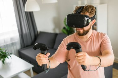 VR Oculus 'u olan sakallı adam oyun deneyimiyle heyecan verici oyun deneyimi olan VR joystick kontrolörleri oturma odasının önünde duruyor. Sanal gerçeklik oyunu konsepti