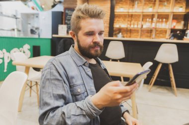 Bearded male using smartphone at modern coffee shop and chatting online messaging on mobile phone. Social networks and reading blog