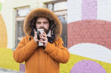 Close-up portrait handsome bearded millennial man in winter clothes and with thermos snow outdoor. Cold season and hot beverage in winter time.