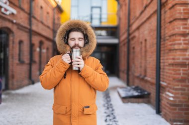 Close-up portrait handsome bearded millennial man in winter clothes and with thermos snow outdoor. Cold season and hot beverage in winter time.