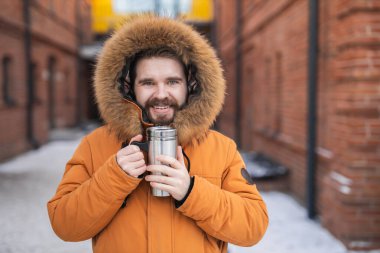 Close-up portrait handsome bearded millennial man in winter clothes and with thermos snow outdoor. Cold season and hot beverage in winter time.