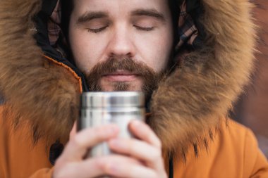 Close-up portrait handsome bearded millennial man in winter clothes and with thermos snow outdoor. Cold season and hot beverage in winter time.