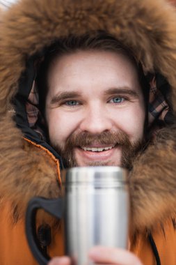 Close-up portrait handsome bearded millennial man in winter clothes and with thermos snow outdoor. Cold season and hot beverage in winter time.