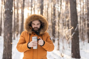 Close-up portrait handsome bearded millennial man in winter clothes and with thermos snow outdoor. Cold season and hot beverage in winter time.