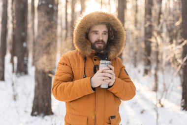 Close-up portrait handsome bearded millennial man in winter clothes and with thermos snow outdoor. Cold season and hot beverage in winter time.