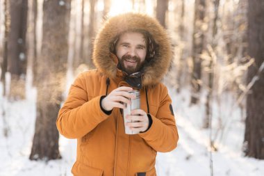 Close-up portrait handsome bearded millennial man in winter clothes and with thermos snow outdoor. Cold season and hot beverage in winter time.