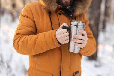 Close-up portrait handsome bearded millennial man in winter clothes and with thermos snow outdoor. Cold season and hot beverage in winter time.