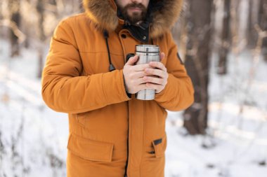 Close-up portrait handsome bearded millennial man in winter clothes and with thermos snow outdoor. Cold season and hot beverage in winter time.