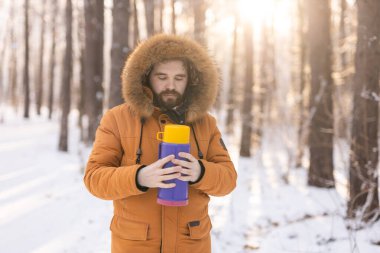 Close-up portrait handsome bearded millennial man in winter clothes and with thermos snow outdoor. Cold season and hot beverage in winter time.