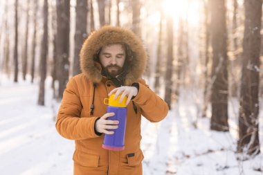 Close-up portrait handsome bearded millennial man in winter clothes and with thermos snow outdoor. Cold season and hot beverage in winter time.