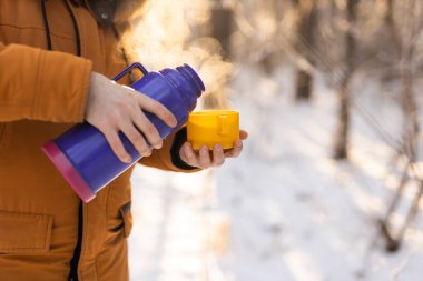 Man pours hot tea from a thermos into a snow walking in snowy frozen winter forest at sunset. Adventure, tourism and camping concept. Copy space and empty place for text advertising.