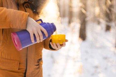 Man pours hot tea from a thermos into a snow walking in snowy frozen winter forest at sunset. Adventure, tourism and camping concept. Copy space and empty place for text advertising.
