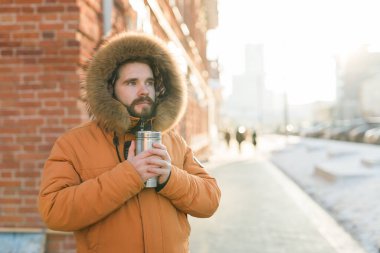 Close-up portrait handsome bearded millennial man in winter clothes and with thermos snow outdoor. Cold season and hot beverage in winter time.