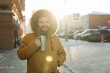 Close-up portrait handsome bearded millennial man in winter clothes and with thermos snow outdoor. Cold season and hot beverage in winter time.
