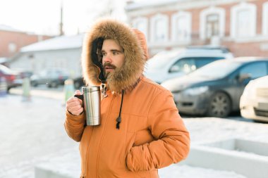 Close-up portrait handsome bearded millennial man in winter clothes and with thermos snow outdoor. Cold season and hot beverage in winter time.