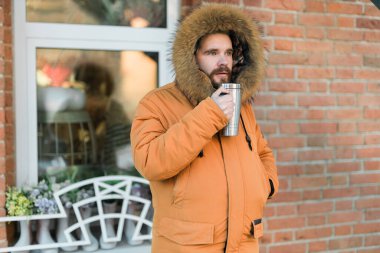 Close-up portrait handsome bearded millennial man in winter clothes and with thermos snow outdoor. Cold season and hot beverage in winter time.