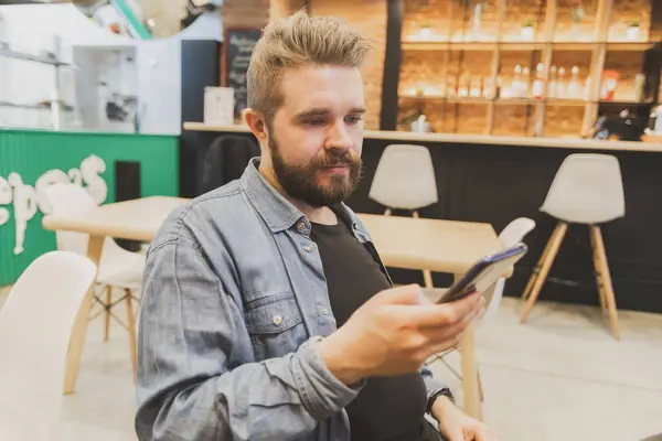 stock image Bearded male using smartphone at modern coffee shop and chatting online messaging on mobile phone. Social networks and reading blog