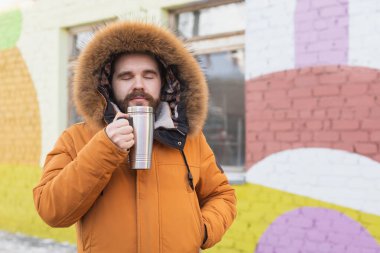 Close-up portrait handsome bearded millennial man in winter clothes and with thermos snow outdoor. Cold season and hot beverage in winter time.