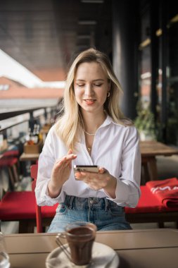 Restoran ve akıllı telefon. Sarışın, yazı yazan, kaydıran ya da e-postayı kontrol eden ya da sosyal medya uygulaması ya da sosyal ağ kullanıcısı ile iletişime geçen bir kadın. Mutlu bir gülümseme ve gezici teknolojisi olan genç bir kız.