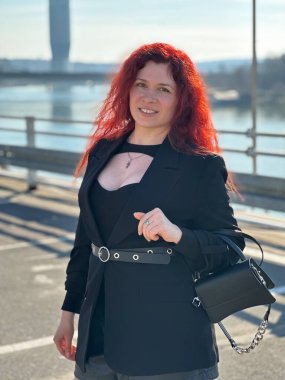 Uzun kıvırcık saçlı, güneşli bir günde kameraya özenle bakan milenyum kadını. Arka planda modern binalar ve bir nehir yer alıyor ve kentte sakinlik hissi uyandırıyor. Şehir hayatı, Belgrad ve