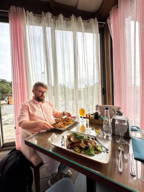 Rahatlamış bir ortamda, dışarısı manzaralı yumuşak pembe perdelerle çevrili cam bir masada yemek yemekten hoşlanan bir adam. Önünde zarif bir şekilde lezzetli bir yemek sunuluyor, bir yemek anını yakalıyor.