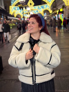 Millennial woman standing outdoors at Christmas night, wrapped in a cozy shearling jacket. the city lights in the background create a warm and Christmas holidays atmosphere, enhancing the peaceful and clipart