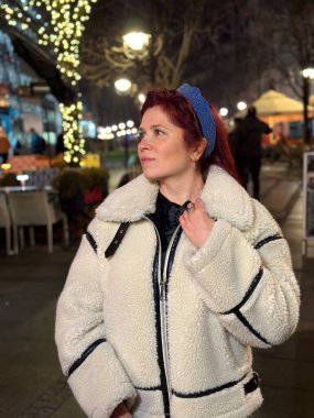 Millennial woman standing outdoors at Christmas night, wrapped in a cozy shearling jacket. the city lights in the background create a warm and Christmas holidays atmosphere, enhancing the peaceful and clipart