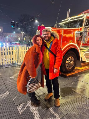 Çift mutlu bir şekilde ışıl ışıl yanan Noel süslü bir kamyonun önünde duruyor ve şehirdeki şenlikli gecenin coşkusunu yakalıyor. Renkli ceketler ve neşeli ifadeler sıcaklık ve sıcaklık yayar.