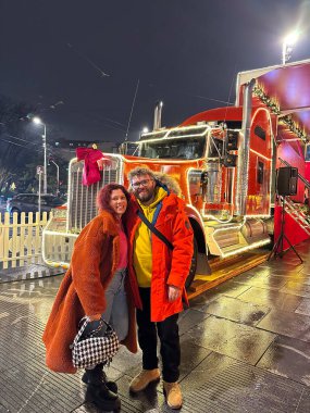 Çift mutlu bir şekilde ışıl ışıl yanan Noel süslü bir kamyonun önünde duruyor ve şehirdeki şenlikli gecenin coşkusunu yakalıyor. Renkli ceketler ve neşeli ifadeler sıcaklık ve sıcaklık yayar.