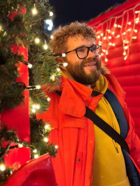 Millennial gülen adam neşeli Noel ışıkları ve dekorasyonlarıyla çevrili, canlı bir tatil ortamında sıcaklık ve neşe saçıyor. Sıcak, tatil ve davetkar atmosfer