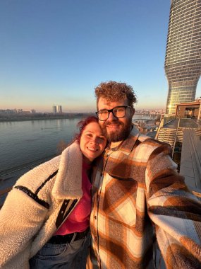 Bir çift balkonda selfie çekiyor, Belgrad 'da günbatımında modern bir gökdeleni olan bir nehre bakıyor. Akşam ışığının sıcacık parıltısı kentsel bir ortam yaratıyor.