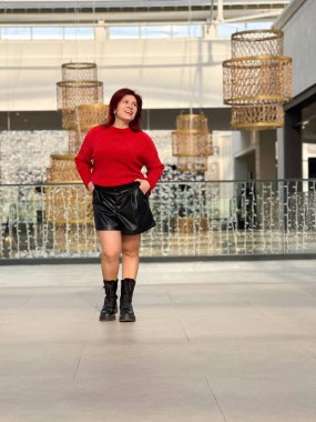 Milenyum kadınları, canlı kırmızı bir kazak ve siyah çizmeler giyerek, açık ve modern bir ortamda güven içinde poz verirler. Neşeli ifade, özgürlük ve bireysellik duygusunu yakalar..