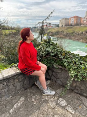 Taştan bir çıkıntıda oturan kırmızı kazaklı, şehir binalarının arka planında huzur dolu nehir manzarasının tadını çıkaran bin yıllık bir kadın. Sonbahar sahnesi kentsel doğanın ve huzurun özünü yakalar.