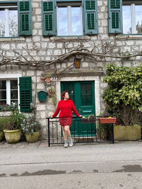 Yeşil kepenkleri olan çekici taş bina ve önünde kırmızı elbiseli kadın duruyor. Kırsal mimarinin ve modanın özünü yansıtan pitoresk bir sahne. Seyahat ve seyahat kavramı.