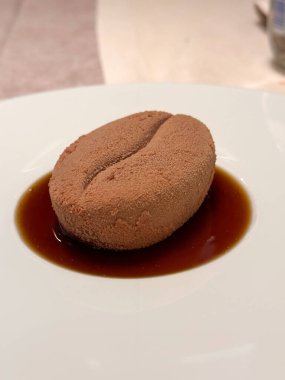 Yakından yapılmış, kahve çekirdeğini andıran bir tatlı tabakta, zengin kahverengi bir sosla çevrili duruyor. Yaratıcı mutfak sunumu, doku ve lezzeti görsel olarak çarpıcı bir şekilde birleştirir.