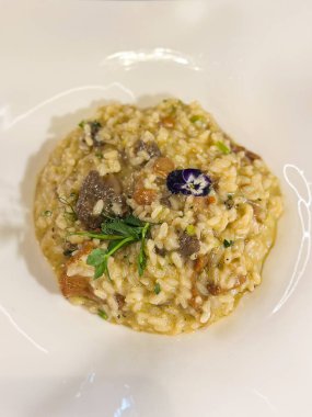 Taze bitkilerle kaplı lezzetli kremalı risotto ve canlı, yenilebilir bir çiçek. Görsel olarak çekici yemekler mutfak sunumunda aroma ve sanatın bir karışımını sergiliyor. Lezzet ve zarafet İtalyanca