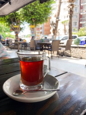 Bir bardak çay kaşıkla beyaz bir fincan çay, arka planda hareketli bir şehir manzarası olan açık bir kafe masasında. Şehir yaşamının ortasında rahatlatıcı bir çay anı..