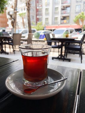 Bir bardak çay kaşıkla beyaz bir fincan çay, arka planda hareketli bir şehir manzarası olan açık bir kafe masasında. Şehir yaşamının ortasında rahatlatıcı bir çay anı..