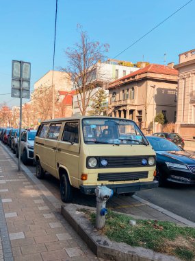 Eski model minibüs, şehir mimarisi ve açık mavi gökyüzüyle çevrili hareketli bir şehir caddesi boyunca park halinde. Manzara, canlı bir mahallede nostalji ve günlük yaşamın bir karışımını yakalar..