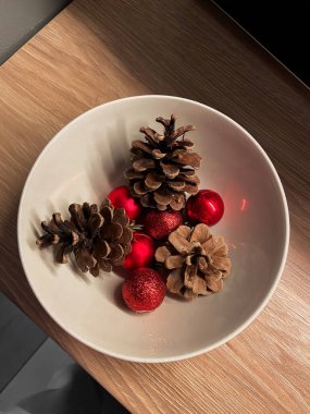 Noel 'de çam kozalakları ve kırmızı süslemelerin sade beyaz bir kasede doğal dokuları ve festival renklerinin altını çizerek düzenlenmesi. Kompozisyon, biraz kırsal dokunuşla mevsimsel dekorun özünü yakalar.