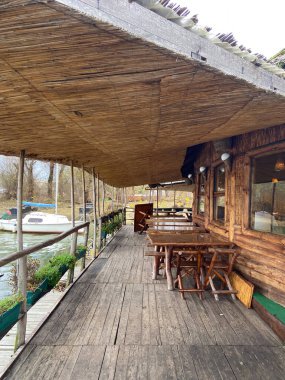 Ahşap masaları ve sakin nehri gören sazdan bir çatısı olan taşralı bir rıhtım güvertesi. Açık hava oturma alanı dinlenmek ve doğal ortamın tadını çıkarmak için sakin bir yer sunar..