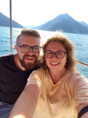 Mutlu çift akıllı telefondan selfie çekiyor ve arka planda çarpıcı dağ manzarası olan manzaralı bir tekne turunun tadını çıkarıyor. Gülümsemeler, suda güzel bir maceranın neşesini ve özgürlüğünü yansıtır.